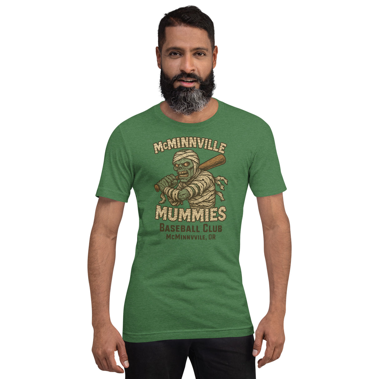 Male model wearing McMinnville Mummies retro baseball t-shirt in heather grass green, showcasing vintage minor league style and spooky mascot.