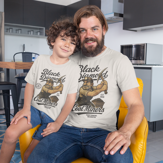 A father and son smiling while wearing matching retro-style Black Diamond Coal Miners t-shirts featuring mascot Dusty McGraw, seated together in a modern kitchen.