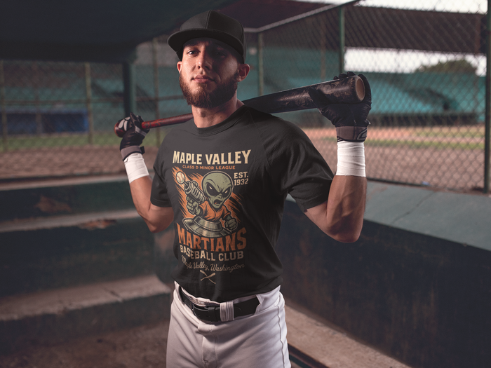 Baseball player standing in a dugout holding a bat over his shoulders, wearing a black Maple Valley Martians t-shirt featuring a vintage alien mascot design and “Class D Minor League” lettering.